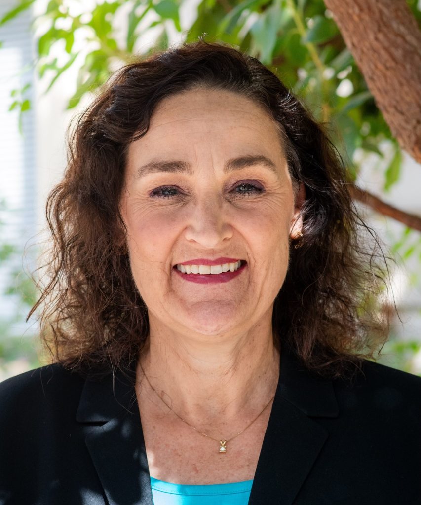 Woman with curly brown hair, wearing a black blazer and blue top, smiling outdoors with green leaves in the background.