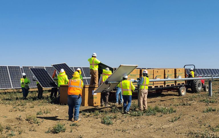 Workers in safety vests install solar panels in a field, unloading equipment from a trailer under a clear blue sky.
