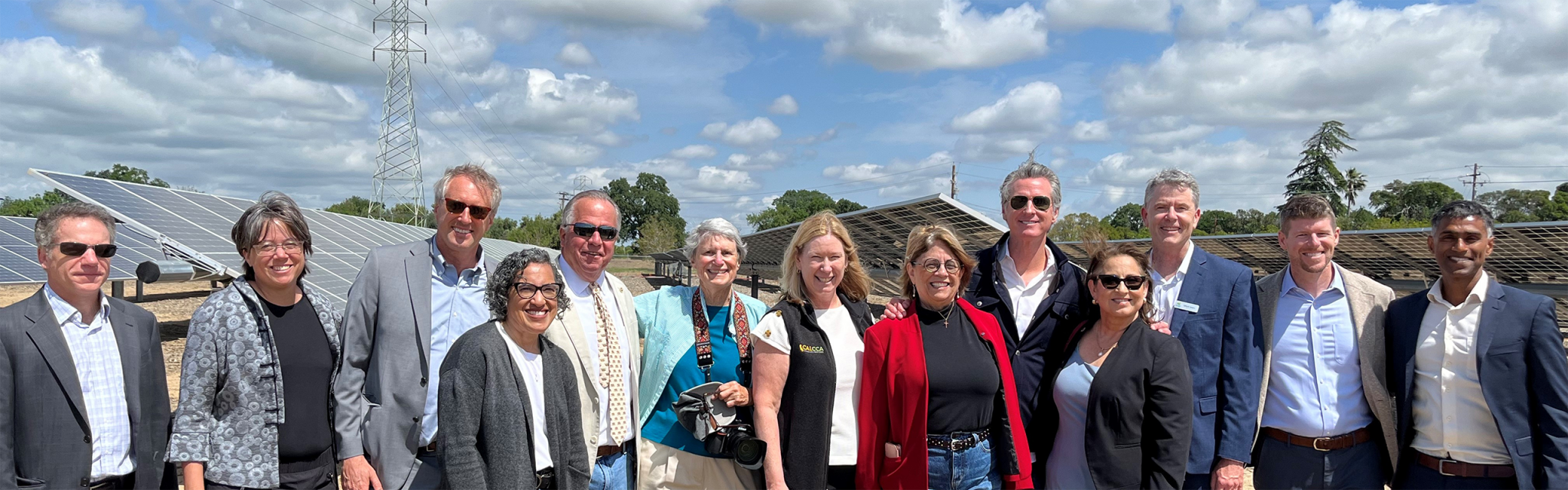 Group of people standing and smiling in front of solar panels outdoors on a sunny day with a partly cloudy sky.