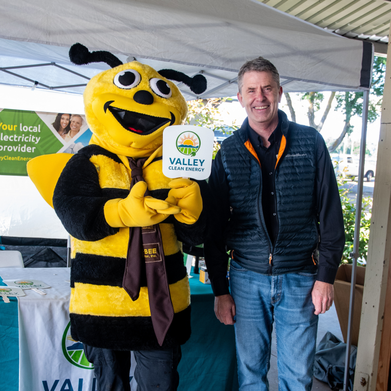 A person stands next to someone in a bee costume holding a Valley Clean Energy sign at an outdoor event booth.