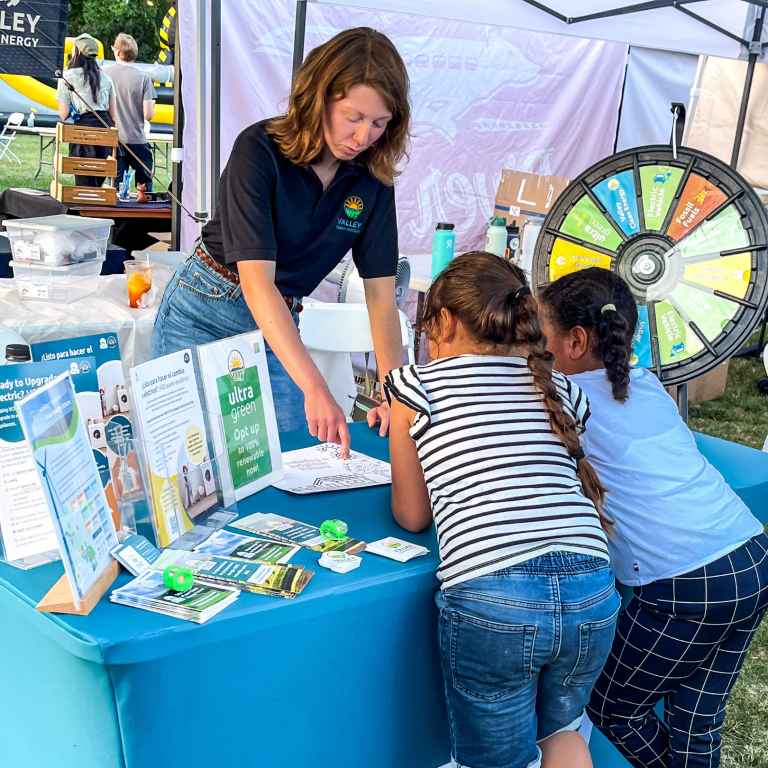 A woman at a booth explains a paper to two girls while a prize wheel and informational displays are visible on the table and in the background.