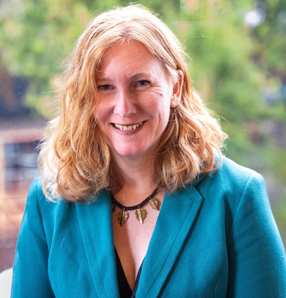 A woman with wavy blonde hair in a teal blazer smiles at the camera, with trees and buildings blurred in the background.