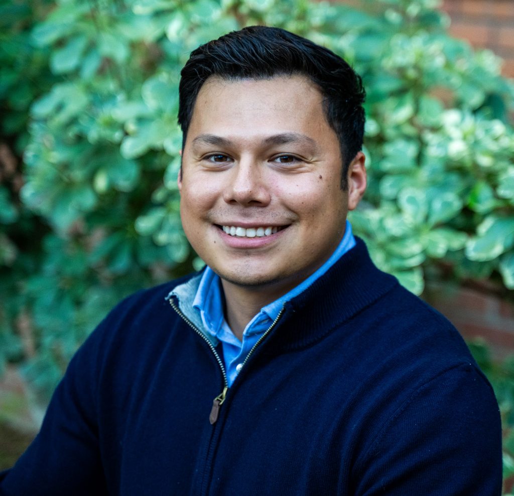 Man with short dark hair wearing a navy sweater over a blue shirt, smiling, sitting outdoors in front of green leafy plants.