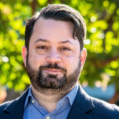 Man with dark hair and beard wearing a blue blazer and shirt stands outdoors with trees in the background.