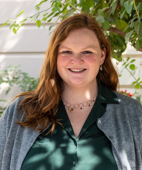 A woman with long red hair, wearing a green shirt and gray cardigan, stands outdoors in front of greenery, smiling at the camera.