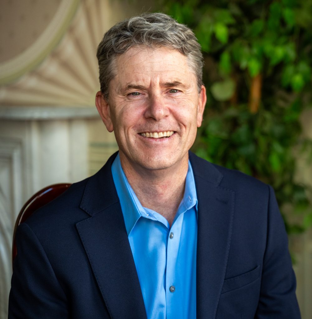 A man in a blue shirt and dark blazer smiles at the camera, seated indoors with greenery in the background.