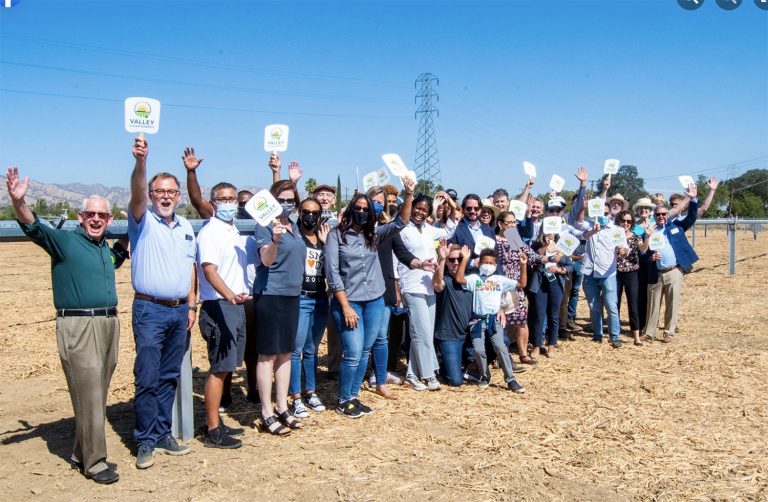A large group of people stand outdoors on straw-covered ground, smiling and holding up small signs, with power lines visible in the background.