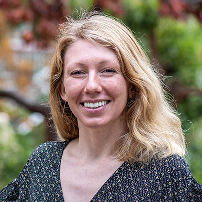 A woman with shoulder-length blonde hair smiles outdoors, wearing a dark patterned top with a blurred green and red background.
