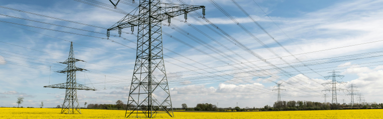 High voltage power lines on metal pylons cross a yellow field under a partly cloudy blue sky.