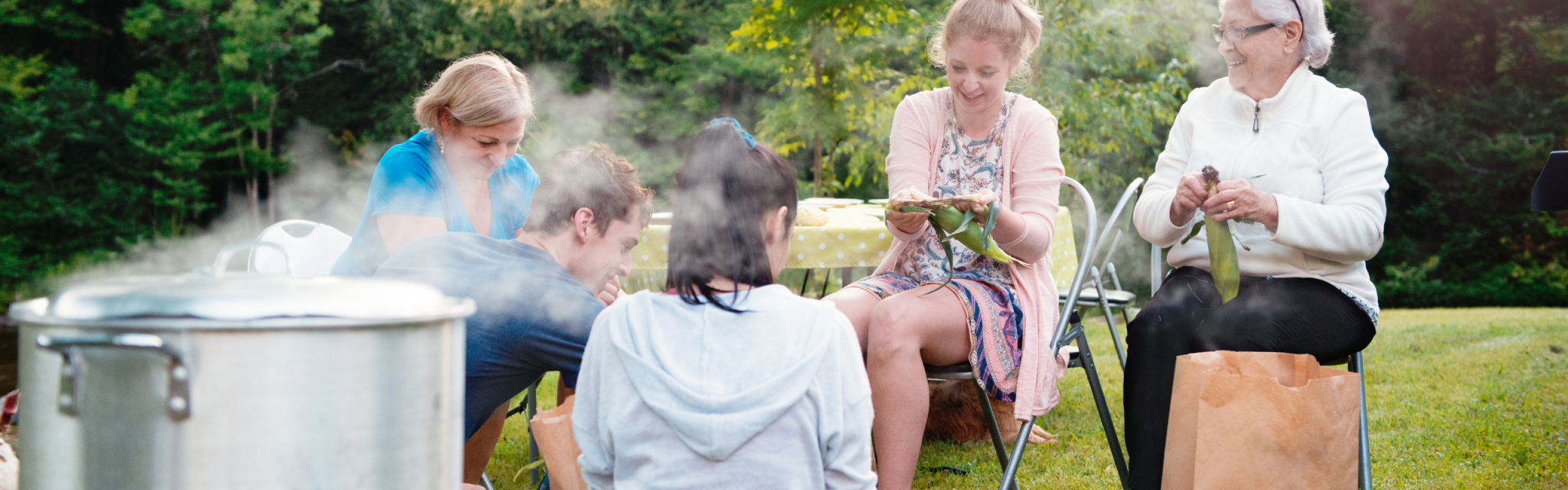 Five people sit outdoors on grass, preparing food together near a steaming pot, with trees in the background.
