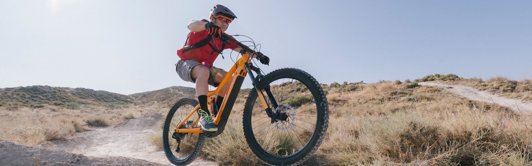 Person wearing a helmet riding a yellow mountain bike off a dirt ramp on a dry, grassy trail under a clear sky.