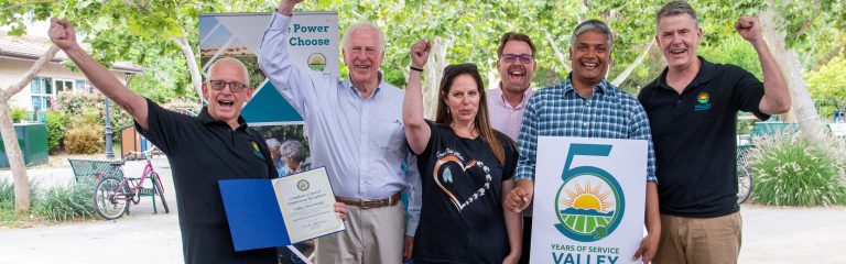 Six adults stand outdoors, some holding a certificate and a “5 Years of Service” sign, all smiling and raising their fists in celebration.