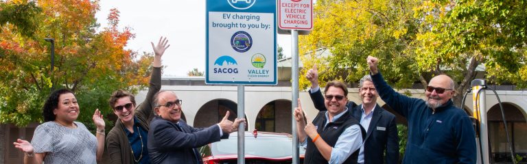 Seis personas posan junto al letrero de una estación de recarga de vehículos eléctricos, sonriendo y levantando las manos, con unos árboles y un edificio al fondo.
