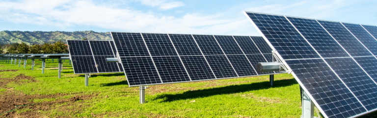 Rows of solar panels installed on a grassy field with mountains and a blue sky in the background.
