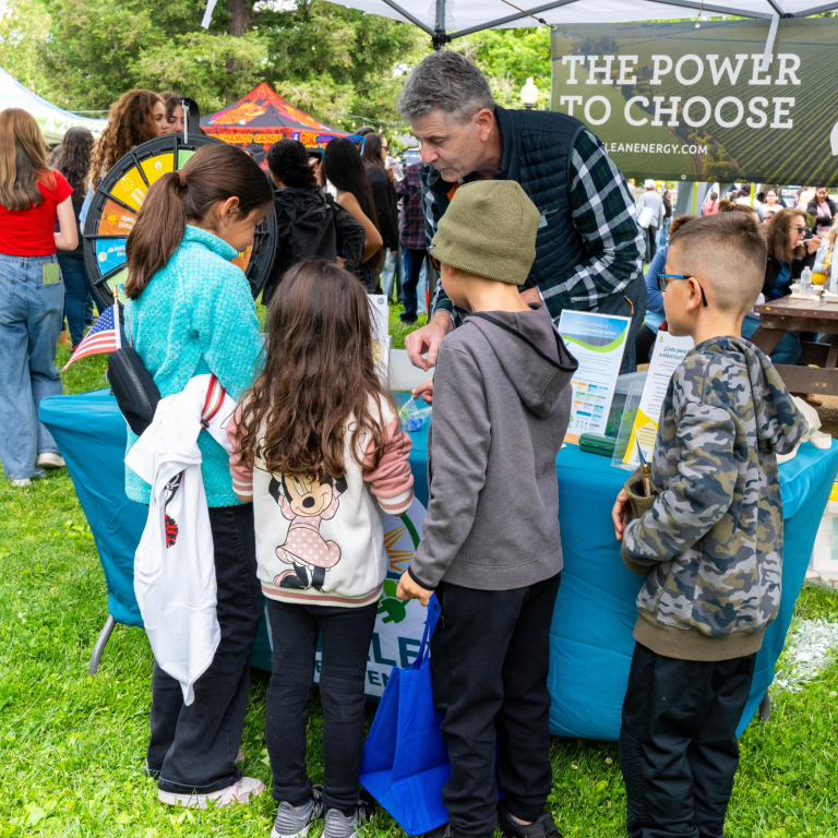 A man talks to four children at an outdoor booth with a sign reading The Power to Choose during a community event.