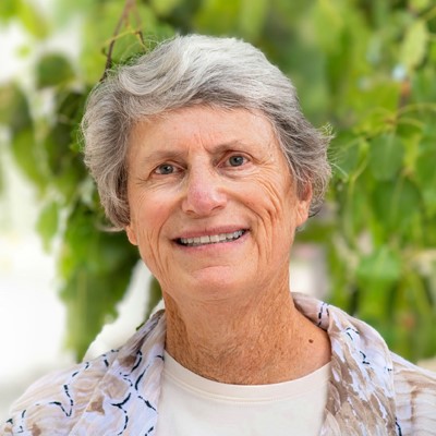 Older woman with short gray hair smiles outdoors in front of green leaves, wearing a light shirt and patterned scarf.