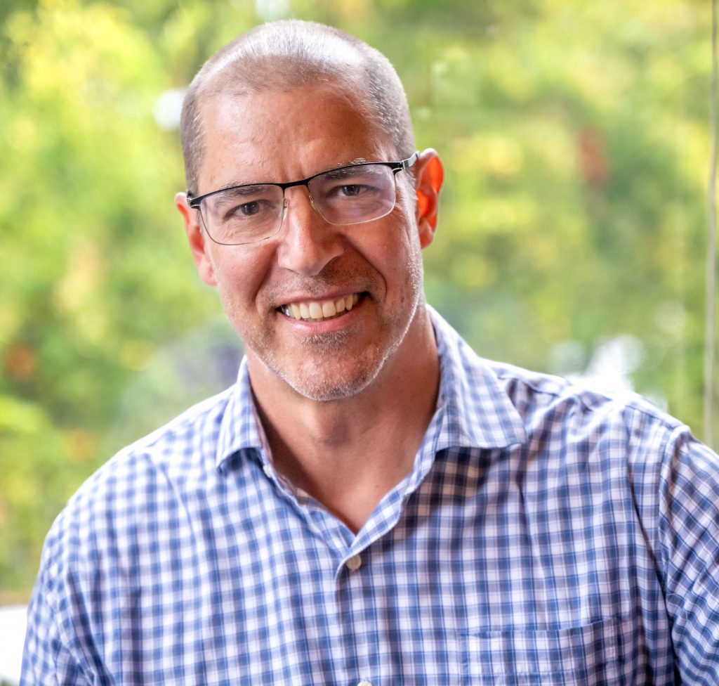 Man wearing glasses and a blue checkered shirt smiles at the camera with a blurred green background.
