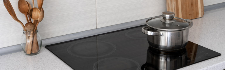 A stainless steel pot with a lid sits on a black electric stovetop next to a jar of wooden utensils and two cutting boards.