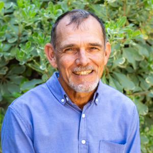 Older man with short hair and goatee, wearing a blue button-up shirt, smiling in front of green leafy plants.