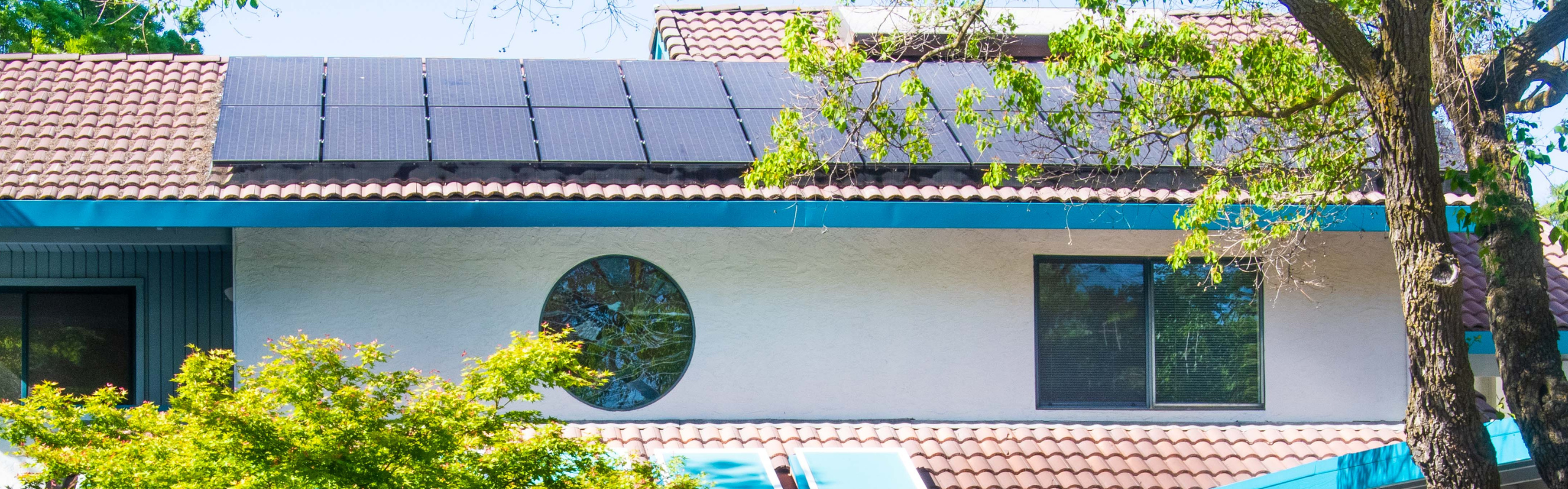 Solar panels are installed on the tiled roof of a residential building with trees in the foreground.