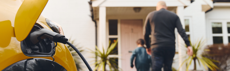 Yellow electric car charging in a driveway while an adult and a child walk toward the entrance of a house in the background.