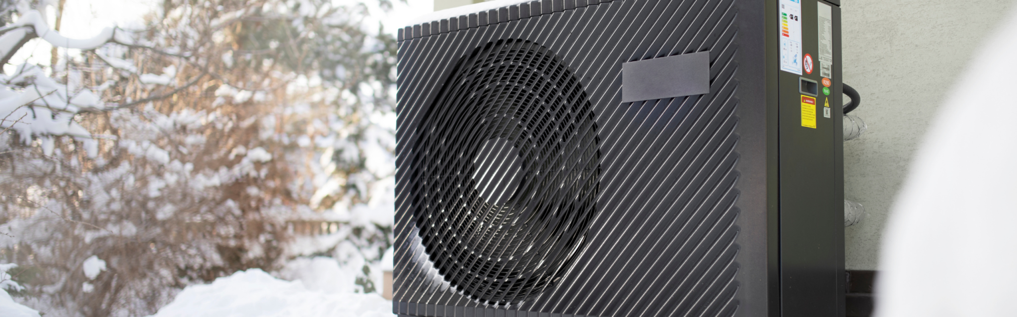 Outdoor heat pump unit in a snowy yard, with trees and a building visible in the background.