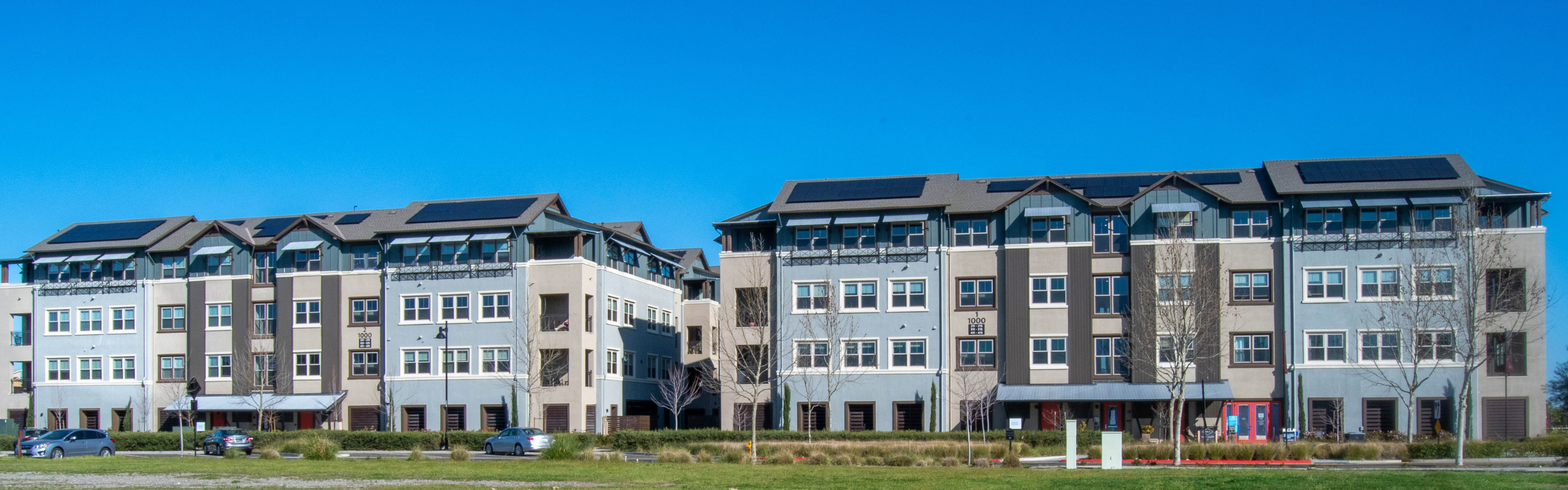 Two modern four-story apartment buildings with solar panels on the roofs, under a clear blue sky.